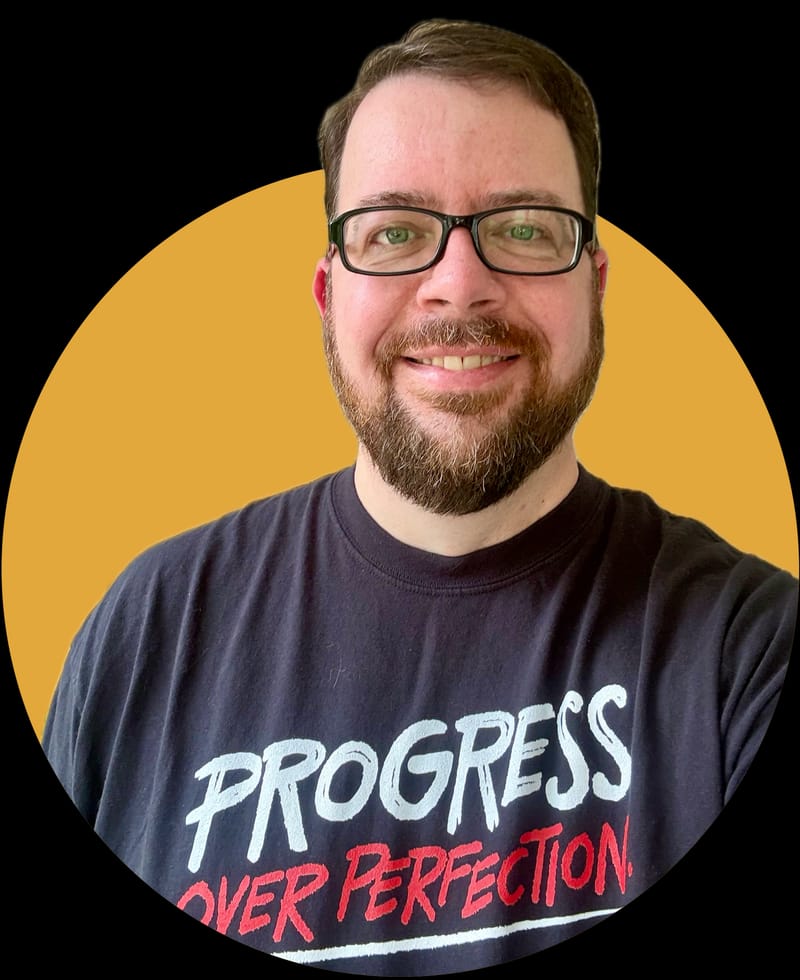 Matt Vanderzalm, a tall white person wearing glasses, smiles at the camera. Matt is wearing a black t-shirt that says Progress over Perfection.